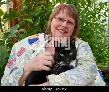 This middle aged woman is smiling and happy as she's holding a black cat with green eyes in a greenhouse. She's wearing glasses Stock Photo