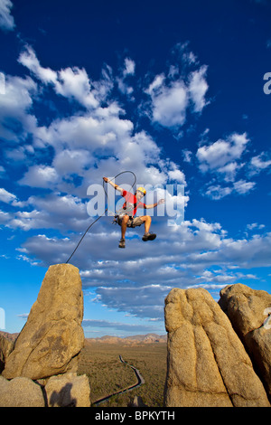 Male rock climber leaps across a gap on the summit of a pinnacle with a cloud filled sky behind him. Stock Photo