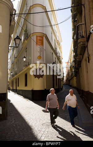 Men walk on typical Spanish cobbled back street / streets / road along with angular buildings and blue sky. Cadiz, Spain. Stock Photo