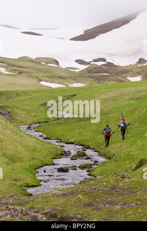 Two skiers hiking up a grassy slope next to a stream on Umnak island in the Aleutian Islands. Stock Photo