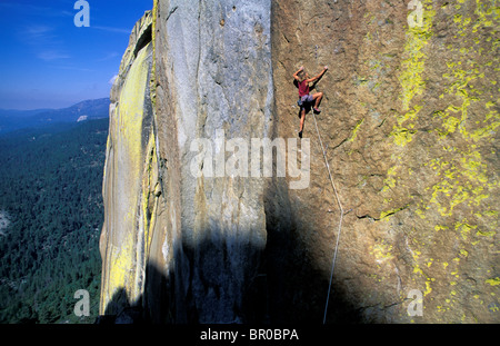 A female rock climber crimping a hold on a cliff face above a forest. Stock Photo