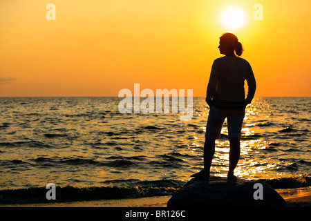 Silhouette of woman standing on a large rock and watching sunset over Lake Winnipeg, Victoria Beach, Manitoba, Canada. Stock Photo