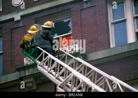 Firefighters / Firemen on Ladder fighting Fire through Broken Window in Apartment Building Stock Photo