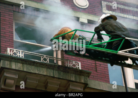 Firefighters / Firemen on Ladder fighting Fire with Hose through Broken Window in Apartment Building Stock Photo