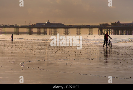 Silhouette of the Palace Pier now called Brighton Pier and cyclist. Low tide at sunset, Brighton. East Sussex, England Stock Photo