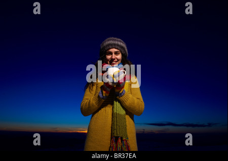 A smiling young woman holds a cup of hot chocolate in front of a sunset sky Stock Photo