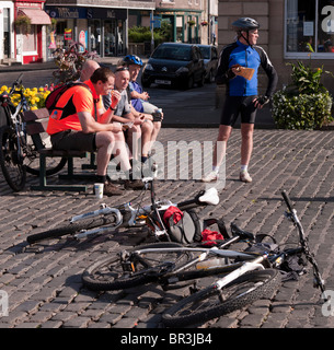 Touring cyclists drop their bikes on the cobbles and sit in the town square to take a break and eat - Scotland, Autumn Stock Photo