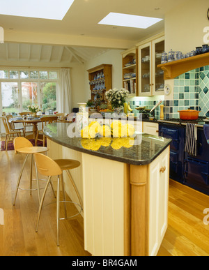 Plywood+stainless-steel stools at breakfast bar on island unit with yellow flowers on granite worktop in large kitchen extension Stock Photo