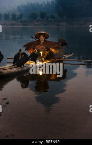 Fisherman wearing straw coat fishing with cormorants on bamboo raft on Li River at dusk, Yangshuo, Guangxi, China Stock Photo