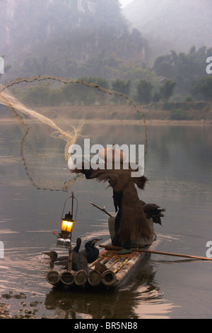Fisherman wearing straw coat with cormorants on bamboo raft casting fishing net on Li River at dusk, Yangshuo, Guangxi, China Stock Photo