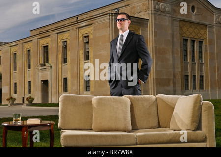 Wealthy young man in suit standing behind a couch and table on the lawn at an art deco mansion Stock Photo