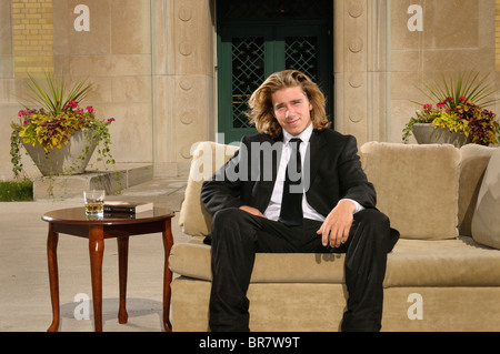 Wealthy young man with long blond hair in suit sitting on a couch on an outdoor terrace at a mansion Stock Photo
