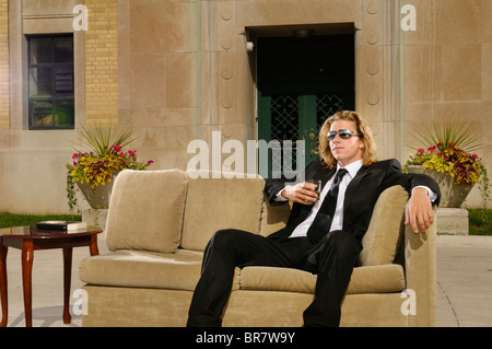 Wealthy young man with long blond hair in suit sitting on a couch with a drink on an outdoor terrace at a mansion Stock Photo