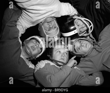 1960s FIVE BOYS IN HUDDLE WEARING HELMETS & FOOTBALL JERSEYS THE VIEW IS FROM INSIDE THE HUDDLE LOOKING UP Stock Photo