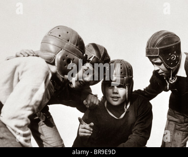 1930s GROUP OF BOYS IN HUDDLE PLAYING FOOTBALL WEARING LEATHER HELMETS Stock Photo