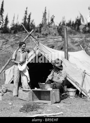 1930s SMILING TALKING COUPLE WORKING BY RUSTIC WESTERN CAMPSITE TENT MAN IN COWBOY HAT SMOKING PIPE WASHING SKILLET WOMAN Stock Photo