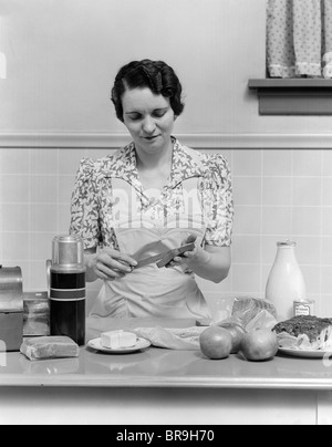 1930s WOMAN HOUSEWIFE IN KITCHEN WEARING APRON MAKING SANDWICH PACKING METAL LUNCH BOX THERMOS APPLES BREAD MILK ON COUNTER Stock Photo