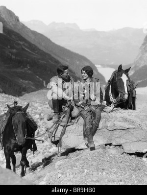 1920s 1930s COUPLE MAN WOMAN WEARING RIDING GEAR JODHPURS BOOTS SPURS SITTING ON LARGE ROCK BY TWO HORSES WITH WESTERN SADDLES Stock Photo