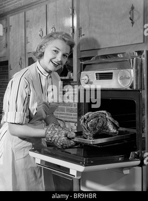 1950s SMILING WOMAN HOUSEWIFE WEARING APRON TAKING LARGE ROAST BEEF FROM ELECTRIC OVEN IN KITCHEN LOOKING AT CAMERA Stock Photo