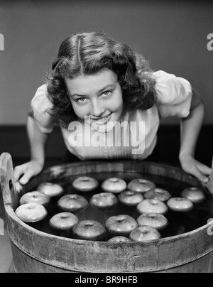 1940s SMILING TEEN GIRL LEANING OVER TUB ABOUT TO BEGIN BOBBING FOR APPLES FLOATING IN THE WATER Stock Photo