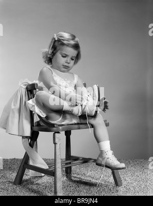 1960s LITTLE GIRL PUTTING ON SHOES SITTING ON CHAIR Stock Photo