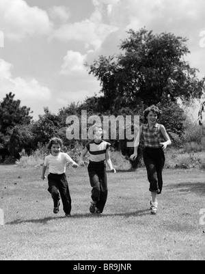 1950s THREE GIRLS RUNNING IN GRASSY FIELD Stock Photo