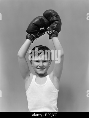 1940s BOY CHILD WINNER WEARING BOXING GLOVES HOLDING HANDS ABOVE HEAD LOOKING AT CAMERA Stock Photo