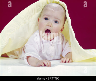 1960s BABY MOUTH WIDE OPEN PEEKING OUT FROM UNDER YELLOW BLANKET LOOKING AT CAMERA Stock Photo