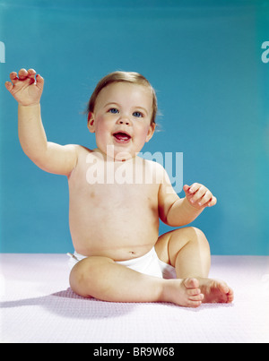 1960s HAPPY SMILING BABY WEARING DIAPER SITTING AND WAVING RAISED ARM LOOKING AT CAMERA Stock Photo