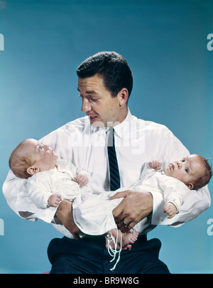 1960s FATHER HOLDING TWIN BABIES INFANTS LOOKING DOWN AT ONE OF THEM Stock Photo