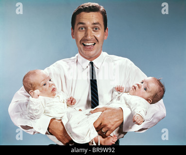 1960s SMILING MAN FATHER LOOKING AT CAMERA HOLDING TWIN BABIES INFANTS Stock Photo