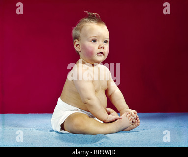 1960s BABY IN DIAPER SITTING UP AGAINST RED BACKGROUND Stock Photo
