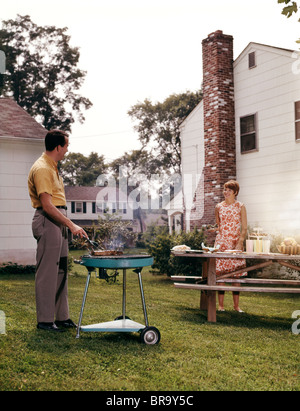 1960s COUPLE SUBURBAN BACKYARD MAN GRILLING WOMAN SETTING PICNIC TABLE Stock Photo