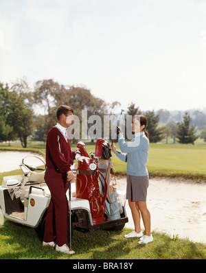 1960s SMILING COUPLE MAN WOMAN SELECTING GOLF CLUBS FROM GOLF BAG ON CART BY SAND TRAP Stock Photo