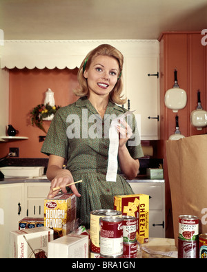 1960s YOUNG BLOND HOUSEWIFE SMILING WHILE CHECKING GROCERY SHOPPING RECEIPT IN KITCHEN Stock Photo