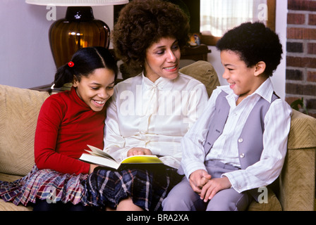 1970 1970s 1980 1980s RETRO AFRICAN AMERICAN MOTHER SITTING ON COUCH WITH SON AND DAUGHTER READING TOGETHER Stock Photo