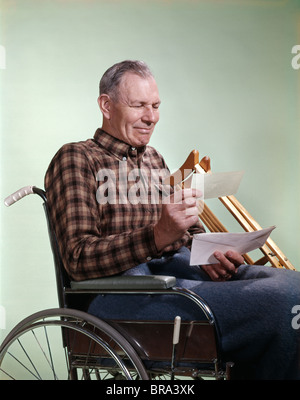 1960 1960s RETRO SMILING SENIOR MAN SITTING IN WHEELCHAIR WITH CRUTCHES READING LETTER INSURANCE CHECK Stock Photo