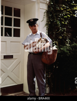 1970s MIDDLE AGED MAILMAN STANDING BY WHITE DOOR SORTING MAIL FOR DELIVERY Stock Photo