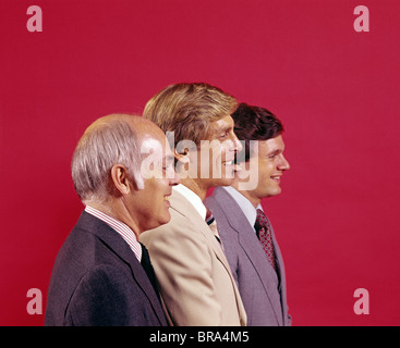 1970 1970s RETRO PORTRAIT PROFILE HEAD AND SHOULDERS OF THREE BUSINESSMEN IN SUITS SMILING RED BACKGROUND Stock Photo