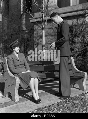 1940s COUPLE MAN IN ARMY UNIFORM TAKING PHOTO OF WOMAN SITTING ON BENCH WEARING HAT Stock Photo