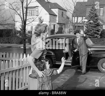 1950s MOTHER AND DAUGHTER WAVING TO FATHER OPENING AUTOMOBILE DOOR IN FRONT OF SUBURBAN HOME Stock Photo