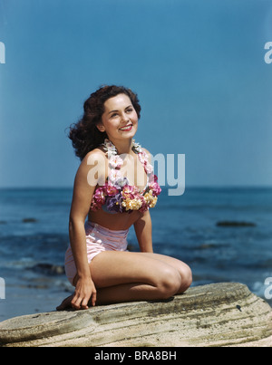 1940s 1950s SMILING BRUNETTE WOMAN SITTING ON ROCK WEARING TWO PIECE BATHING SUIT APPLIQUÉD WITH PASTEL FLOWERS Stock Photo