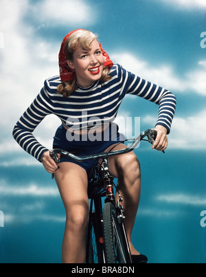 1940s 1950s SMILING BLOND WOMAN RIDING BICYCLE WEARING RED BANDANA BLUE AND WHITE STRIPED SHIRT Stock Photo