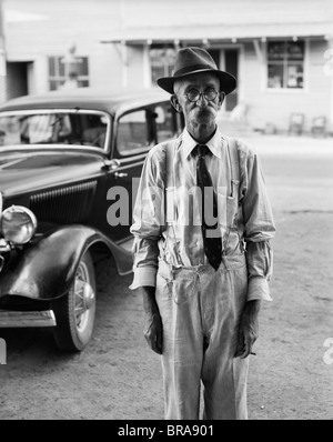 1930s PORTRAIT OF OLD MAN WEARING HAT GLASSES TIE & SUSPENDERS CAR IN BACKGROUND Stock Photo
