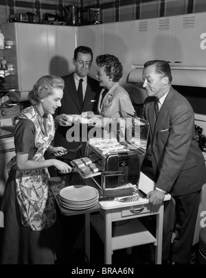 1950s 2 COUPLES IN KITCHEN WOMAN COOKING HAM & EGGS IN CHROME BROILER Stock Photo