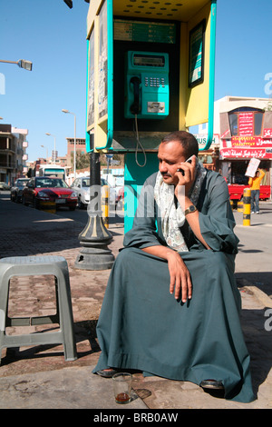 Man sitting under a public telephone using a mobile phone. The latter has replaced the former, Hurghada, Egypt Stock Photo