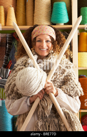 Woman Wearing Knitted Scarf Standing In Front Of Yarn Display Holding Giant Needles And Ball Of Wool Stock Photo