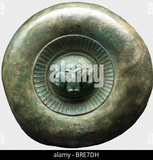 A decorative Etruscan shield, circa 500 B.C. Bronze. The separately made central medallion displays a ram's head in high relief with remnants of white eye inlays (colour paint?), surrounded by an ornamental ring bearing a tongue pattern, and a broad, strongly rounded protuding rim. Diameter 28.5 cm. Broken into small pieces, glued together and stabilised with fiberglass. Restorations at various places. Axel Guttmann Collection (R 203/AG 195). Shield-shaped plates of this kind appear in great quantities in Etruscan graves and are generally decorated with a lion , Stock Photo