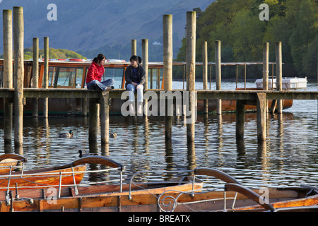 Two girls chatting while seated on the landing stage by the rowing boats on Derwentwater, Keswick Stock Photo
