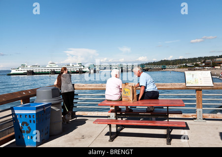 senior couple perched on top of picnic table enjoy  brown bag picnic as dogwalker looks at view & ferry leaves pier in distance Stock Photo